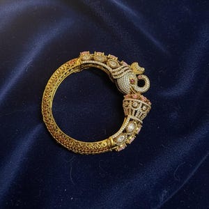 May include: Gold-toned bangle bracelet with an elephant head design. The bracelet features intricate detailing with white and pink gemstones. The elephant head is adorned with white stones and a red accent. The bracelet is set against a dark blue background.