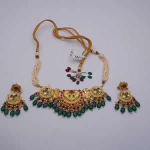 May include: A gold-coloured necklace and matching earrings set. The necklace features a central pendant with red, green, and white accents, and is strung with a cream-coloured cord. The earrings have a similar design, with green teardrop-shaped beads.