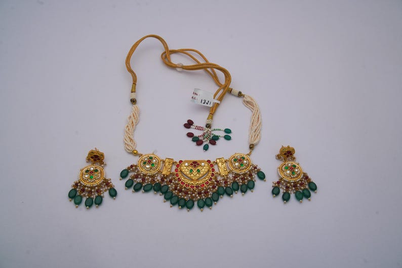 May include: A gold-coloured necklace and matching earrings set. The necklace features a central pendant with red, green, and white accents, and is strung with a cream-coloured cord. The earrings have a similar design, with green teardrop-shaped beads.