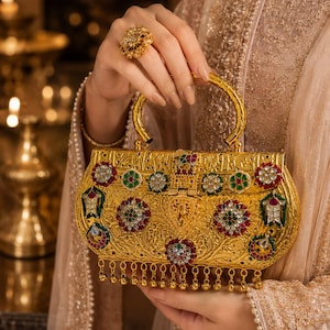 May include: A gold-toned metal handbag featuring intricate embossed floral patterns, inlaid with colorful red, green, and white stones. The bag has a curved handle, a hinged clasp, and a row of small dangling gold beads along the bottom edge.