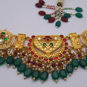 May include: Gold necklace with a heart-shaped pendant, red and green detailing, and dangling green beads. The necklace has intricate detailing, pearls, and a beaded strand.