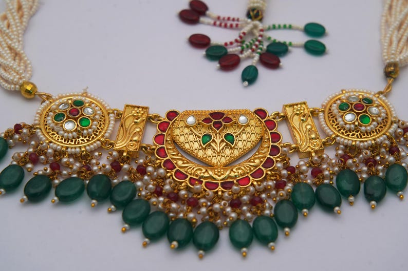 May include: Gold necklace with a heart-shaped pendant, red and green detailing, and dangling green beads. The necklace has intricate detailing, pearls, and a beaded strand.