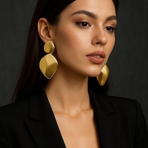 Puede incluir: Pendientes dorados llamativos con un diseño de dos partes. Los pendientes tienen un acabado liso y mate y una forma geométrica. La modelo lleva una blazer negra. El fondo es gris oscuro.