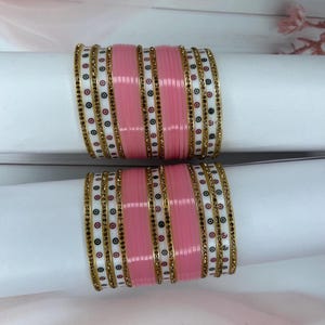 May include: Two sets of pink and white bangles with gold detailing. The bangles feature a repeating pattern of pink, white, and gold bands, with small black and gold embellishments. They are presented on white cylindrical supports.