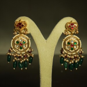 May include: Elaborate gold earrings with detailed design. Each earring has a heart-shaped top with red and gold elements, a circular centre with red, green, and white stones, and hanging green beads. Small pearls and red beads provide additional decoration.