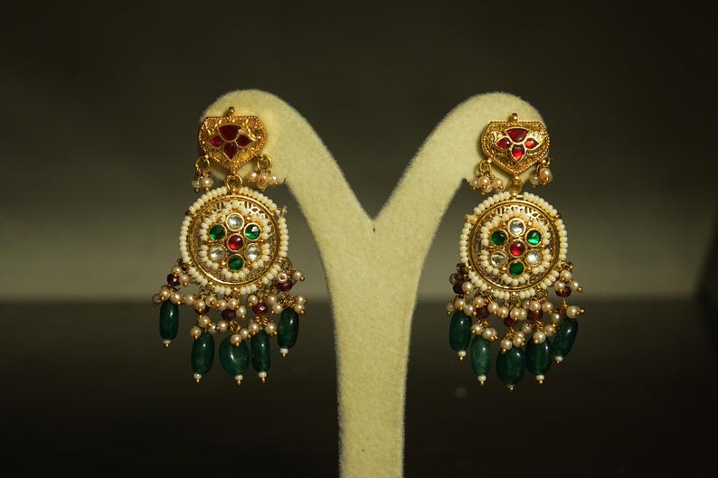 May include: Elaborate gold earrings with detailed design. Each earring has a heart-shaped top with red and gold elements, a circular centre with red, green, and white stones, and hanging green beads. Small pearls and red beads provide additional decoration.