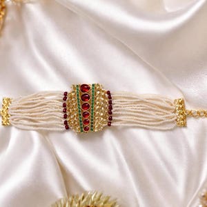 May include: An ornate bracelet featuring multiple strands of off-white beads. The centerpiece is a gold-toned rectangular element adorned with red and green gemstones. The bracelet is set against a white, silky background.