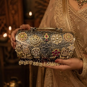 Pode incluir: Embreagem de prata ornamentada com intrincados desenhos florais e geométricos, adornada com pedras preciosas coloridas e pérolas. A bolsa apresenta uma alça superior e um fecho decorativo, mostrando uma estética vintage.