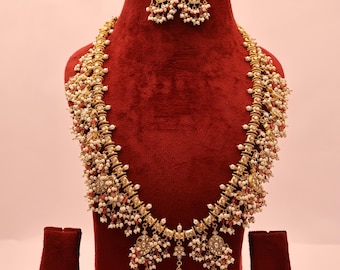 May include: A gold necklace and earring set on a red velvet bust. The necklace has white and red beads, with a large pendant. The earrings have a matching design. A beautiful jewellery set.