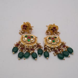 May include: A pair of gold earrings with detailed design. Each earring has a floral top with red accents, a circular centre with green, red, and white stones, and hanging green teardrop beads. Small white pearls are also included in the design.