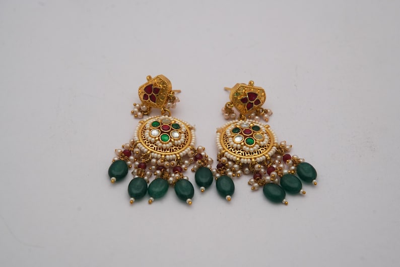 May include: A pair of gold earrings with detailed design. Each earring has a floral top with red accents, a circular centre with green, red, and white stones, and hanging green teardrop beads. Small white pearls are also included in the design.