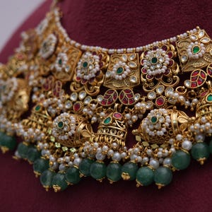 Pu&ograve; includere: Una collana d'oro con dettagli intricati, con pietre preziose verdi e rosse, perle bianche e campanelle pendenti. La collana &egrave; decorata con un motivo floreale e ha uno stile tradizionale indiano.