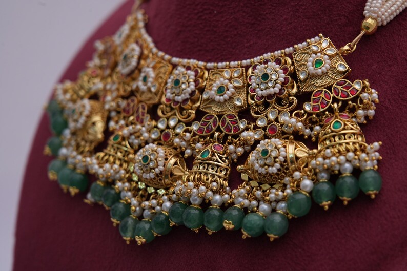 Pu&ograve; includere: Una collana d'oro con dettagli intricati, con pietre preziose verdi e rosse, perle bianche e campanelle pendenti. La collana &egrave; decorata con un motivo floreale e ha uno stile tradizionale indiano.