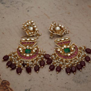 Può includere: Un paio di orecchini dorati ornati. Ogni orecchino presenta una pietra verde centrale, circondata da un design rosa e oro, piccole perle bianche e perline pendenti color bordeaux. Gli orecchini hanno un'estetica vintage e dettagliata.
