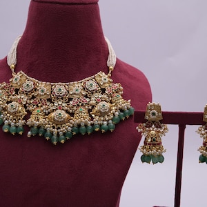 Pu&ograve; includere: Una collana d'oro con pietre verdi e rosse e orecchini abbinati. La collana ha un grande ciondolo decorato con un motivo floreale. Anche gli orecchini sono floreali e hanno un design pendente.