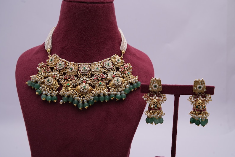 Pu&ograve; includere: Una collana d'oro con pietre verdi e rosse e orecchini abbinati. La collana ha un grande ciondolo decorato con un motivo floreale. Anche gli orecchini sono floreali e hanno un design pendente.