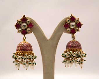 May include: A pair of gold-toned earrings with a floral design at the top, featuring red and clear accents. The earrings have a bell-shaped drop with a red, textured surface, and are adorned with white and green beads.