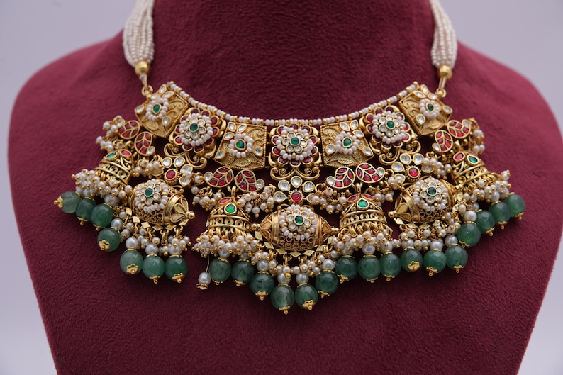 Pu&ograve; includere: Una collana d'oro con un grande ciondolo ornato con pietre rosse, verdi e bianche. Il ciondolo &egrave; circondato da piccole perle perlate e pietre verdi.