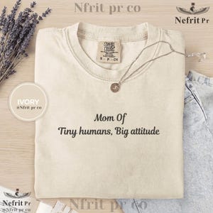以下が含まれることがあります： 「Mom Of Tiny humans, Big attitude」の黒い文字がプリントされたアイボリー色のTシャツ。ペンダント付きのシルバーネックレスがシャツの上にあります。シャツにはComfort Colorsのタグが付いています。