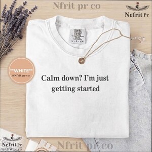 May include: White t-shirt with the text "Calm down? I'm just getting started" in black. The shirt has a crew neck and short sleeves. A gold necklace with a pendant is draped across the shirt. The shirt is on a wooden surface.