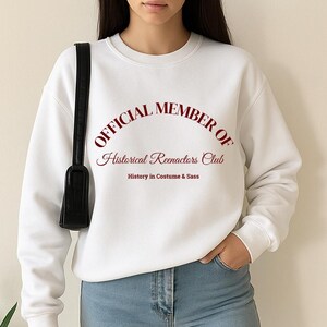 May include: White sweatshirt with the text "OFFICIAL MEMBER OF Historical Reenactors Club" in burgundy script. The sweatshirt also has the text "History in Costume & Sass" below the club name. The person is wearing blue jeans and a black purse.