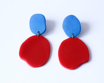 May include: A pair of abstract earrings. Each earring features a small, blue, oval-shaped top piece connected to a larger, red, organic-shaped bottom piece. The earrings are set against a white background.