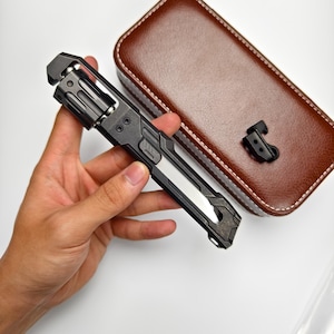May include: A black and silver folding knife with a textured handle is held in a hand. A brown leather case with white stitching and a black clip are in the background. The knife has a silver blade and a complex design.