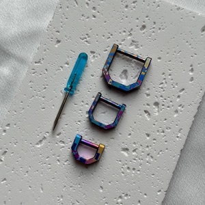 Op de afbeelding: Een assortiment iriserende D-vormige metalen sluitingen in verschillende maten, samen met een kleine blauwe schroevendraaier. De sluitingen hebben een geometrisch patroon met blauw, paars en goud.