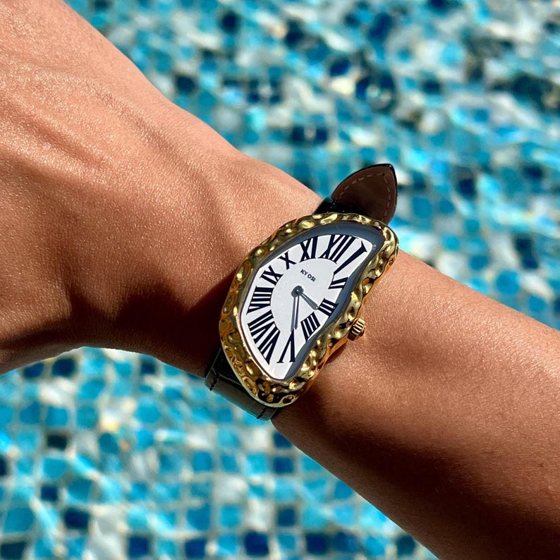 May include: A gold-toned wristwatch with a unique, melted-style face and black Roman numerals. The watch has a white dial and a black leather strap. The watch is on a wrist, with a blurred blue water background.