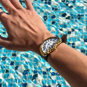 May include: A gold-toned wristwatch with a white face and black Roman numerals. The watch has a unique, textured bezel and a black leather strap. The watch is on a wrist, with a ring on the hand.