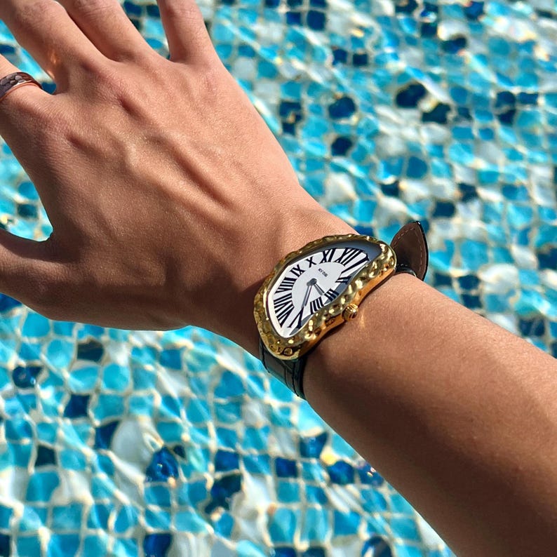 May include: A gold-toned wristwatch with a white face and black Roman numerals. The watch has a unique, textured bezel and a black leather strap. The watch is on a wrist, with a ring on the hand.