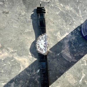 May include: A silver-toned wristwatch with a distorted, melting-style face and black leather band. The watch face features Roman numerals and a white background. The band has a crocodile-like texture.