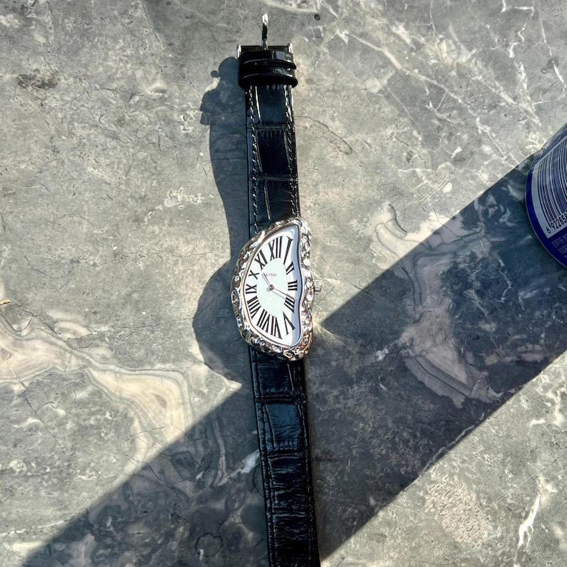 May include: A silver-toned wristwatch with a distorted, melting-style face and black leather band. The watch face features Roman numerals and a white background. The band has a crocodile-like texture.