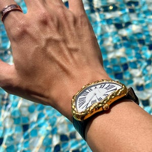 May include: A gold-toned wristwatch with a white face and black Roman numerals. The watch has a unique, irregular shape and a black leather band. A hammered copper ring is also visible.