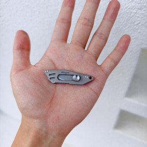May include: A small, silver-colored utility knife with the text "Pioneer EDC" printed on the side. The knife has a rounded blade and a small, circular locking mechanism. The knife is shown in the palm of a hand.