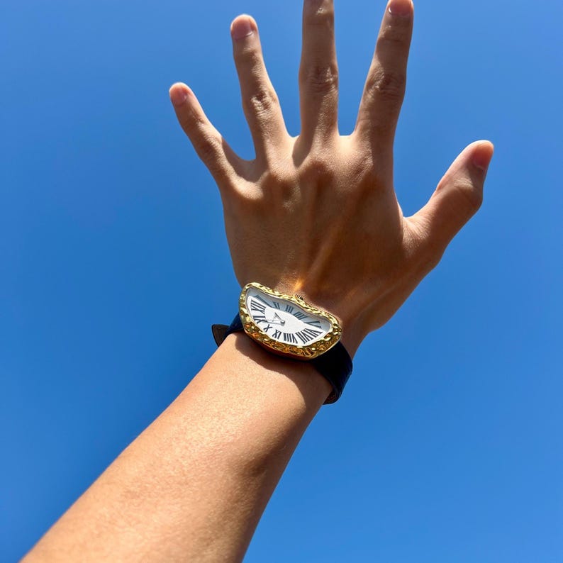 May include: A gold-toned wristwatch with a white face and black leather band. The watch has Roman numeral hour markers and a unique, melted design. The background is a clear, bright blue sky.
