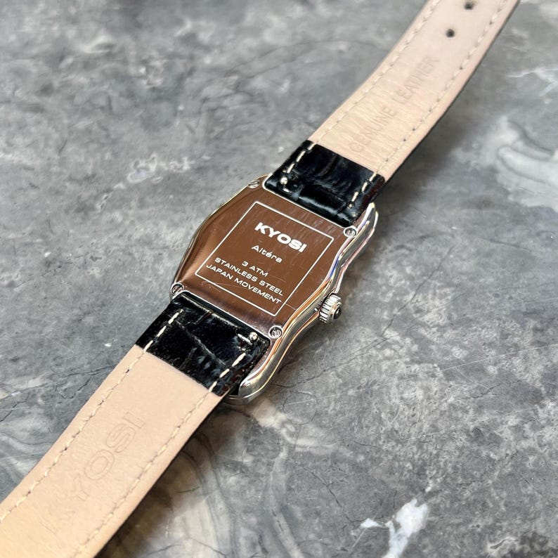 May include: A silver-toned KYOSI Alt&egrave;re watch with a black leather band. The watch face is rectangular with rounded edges. The back of the watch has text that reads "3 ATM Stainless Steel Japan Movement". The watch band is light beige.