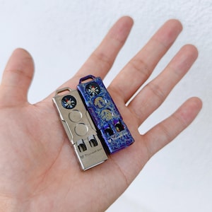 May include: Two small, rectangular metal tools with compasses and other features. One is silver, the other is purple with gold accents. Both have a loop at the top and the text "Pioneer EDC".