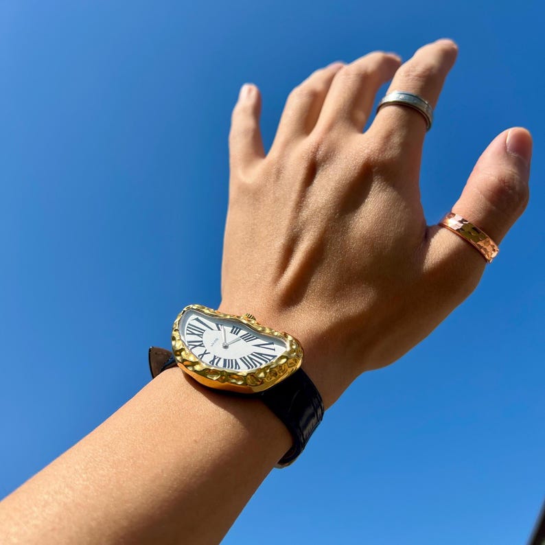May include: A gold-toned wristwatch with a white face and black Roman numerals is worn on a wrist with a black leather band. The watch has a unique, melted shape. Two rings are on the fingers. The background is a clear blue sky.