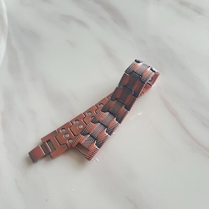 Op de afbeelding: Een koperkleurige armband met een geweven ontwerp en rechthoekige schakels. De armband heeft een magnetische sluiting en is gemaakt van metaal. De armband ligt op een wit oppervlak.