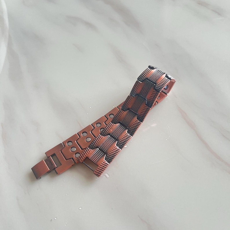 Op de afbeelding: Een koperkleurige armband met een geweven ontwerp en rechthoekige schakels. De armband heeft een magnetische sluiting en is gemaakt van metaal. De armband ligt op een wit oppervlak.
