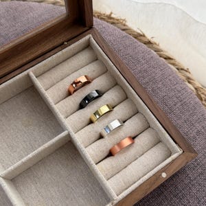 May include: A wooden jewelry box with a glass top, showcasing five rings in various metallic finishes: rose gold, black, gold, silver, and copper. The rings are displayed on a beige fabric insert.