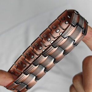 Op de afbeelding: Een koperkleurige armband met een geometrisch ontwerp. De armband is gemaakt van rechthoekige schakels met kleine, cirkelvormige accenten. De schakels zijn verbonden door gestructureerde, patroonsecties. De armband wordt door een hand vastgehouden.