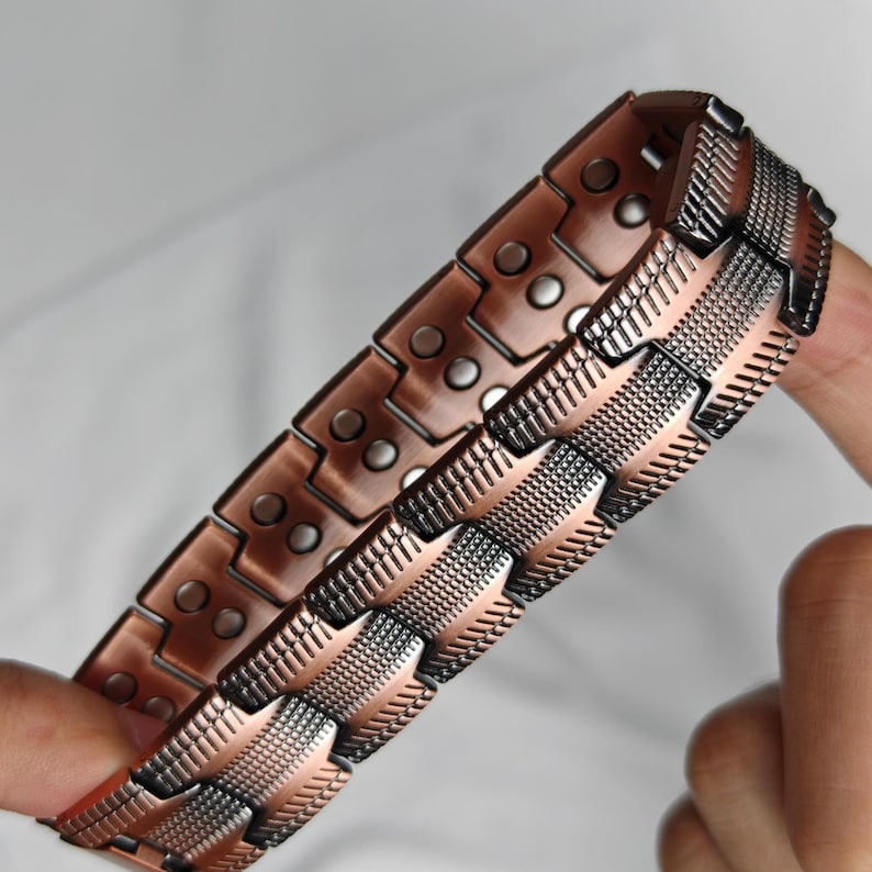 Op de afbeelding: Een koperkleurige armband met een geometrisch ontwerp. De armband is gemaakt van rechthoekige schakels met kleine, cirkelvormige accenten. De schakels zijn verbonden door gestructureerde, patroonsecties. De armband wordt door een hand vastgehouden.