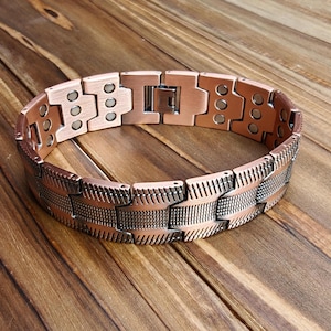 Op de afbeelding: Een koperkleurige armband met een magnetische sluiting. De armband heeft een reeks rechthoekige schakels met een textuurpatroon. De schakels zijn verbonden door kleinere, rechthoekige stukken. De armband wordt gepresenteerd op een houten oppervlak.