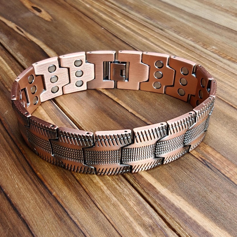 Op de afbeelding: Een koperkleurige armband met een magnetische sluiting. De armband heeft een reeks rechthoekige schakels met een textuurpatroon. De schakels zijn verbonden door kleinere, rechthoekige stukken. De armband wordt gepresenteerd op een houten oppervlak.