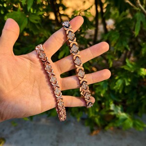 May include: Two rose gold bracelets with a clover and X design, each adorned with small, sparkling crystals. The bracelets are displayed against a backdrop of green foliage, showcasing their elegant design.