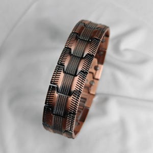 Op de afbeelding: Een koperkleurige armband met een geometrisch ontwerp. De armband heeft een reeks rechthoekige schakels met een getextureerd, donkergrijs patroon. De schakels zijn verbonden door kleinere, koperkleurige stukken. De armband wordt op een witte achtergrond weergegeven.