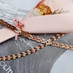 Può includere: Due braccialetti color rame con un design geometrico sono incrociati su una superficie grigia. I braccialetti hanno sezioni alternate di rame massiccio e un motivo intrecciato. Sullo sfondo c'è una composizione floreale di carta rosa.