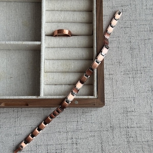 May include: A copper-toned ring and bracelet are displayed in a wooden jewelry box. The ring sits in a compartment, while the bracelet lies diagonally across a textured surface. The bracelet features rectangular links.